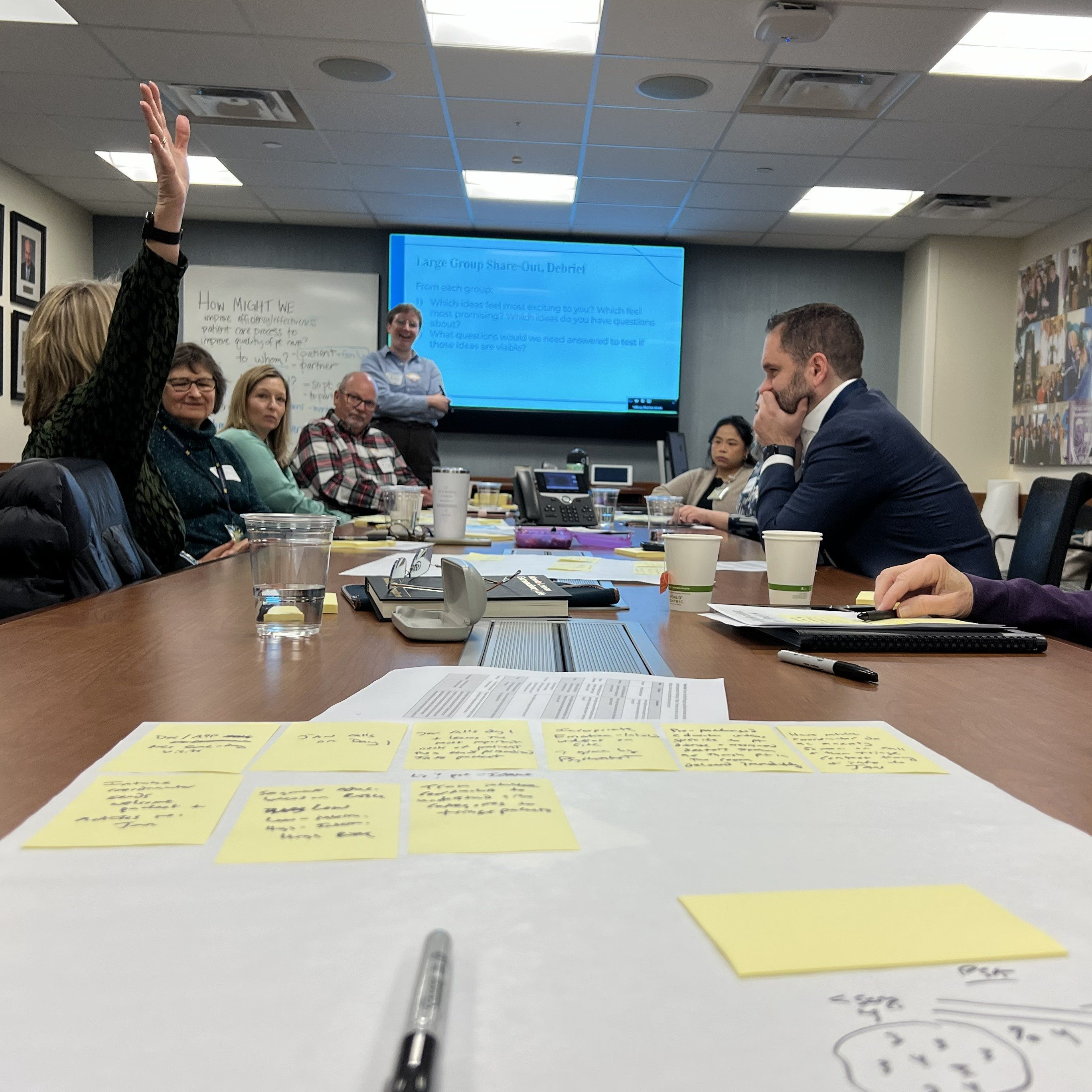 Laura is standing at the front of a large conference room table. A variety of people are sitting around writing ideas on paper and sticky notes; one person has their hand raised.