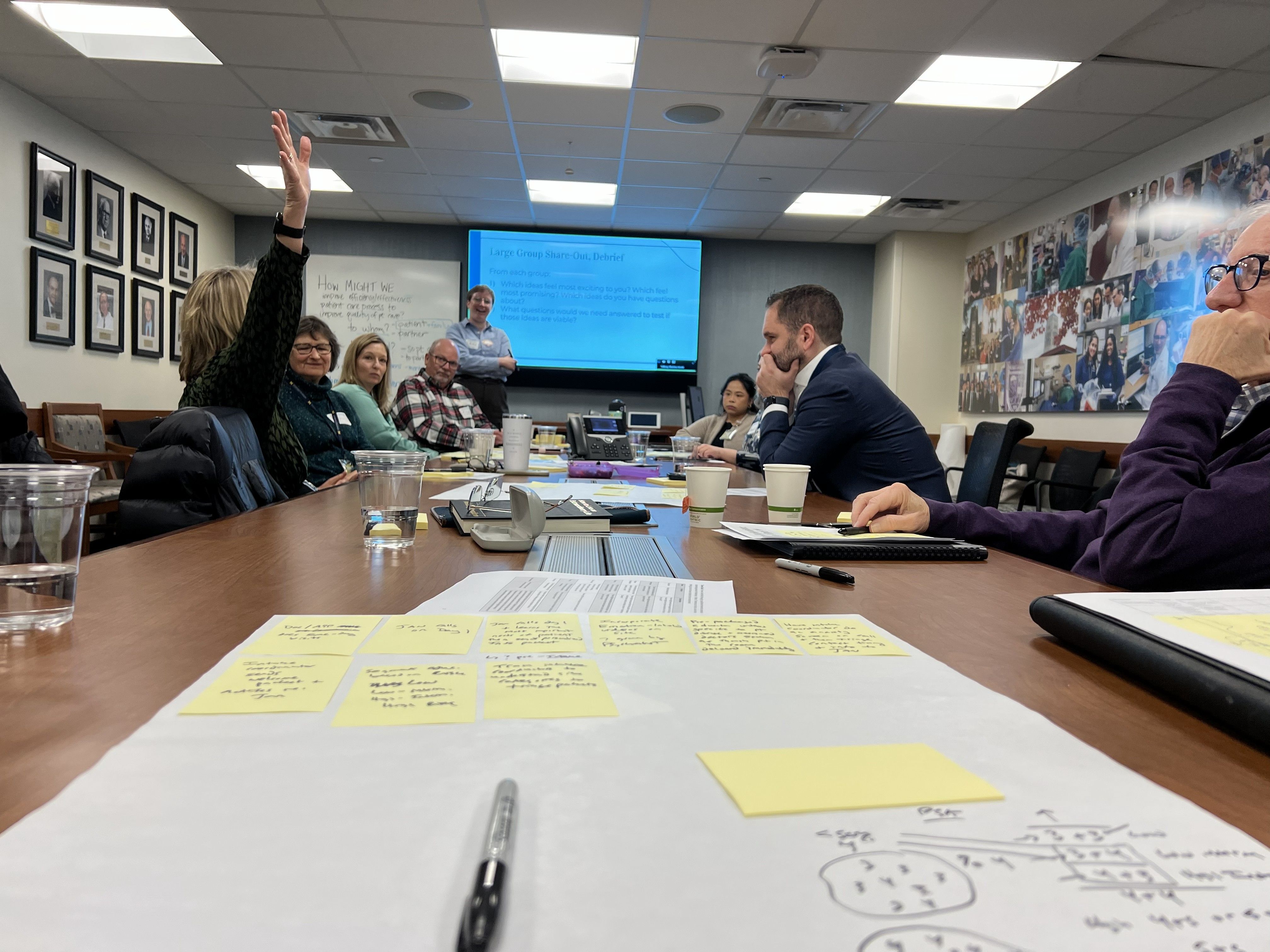 Laura is standing at the front of a large conference room table. A variety of people are sitting around writing ideas on paper and sticky notes; one person has their hand raised.