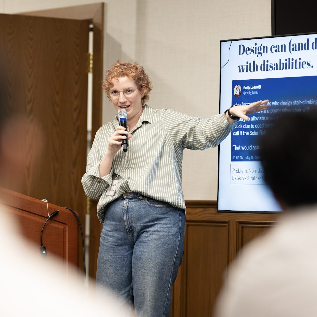 Laura is standing in front of a room full of professionals enthusiastically gesturing at her slides which say "Design can and does harm disabled people."