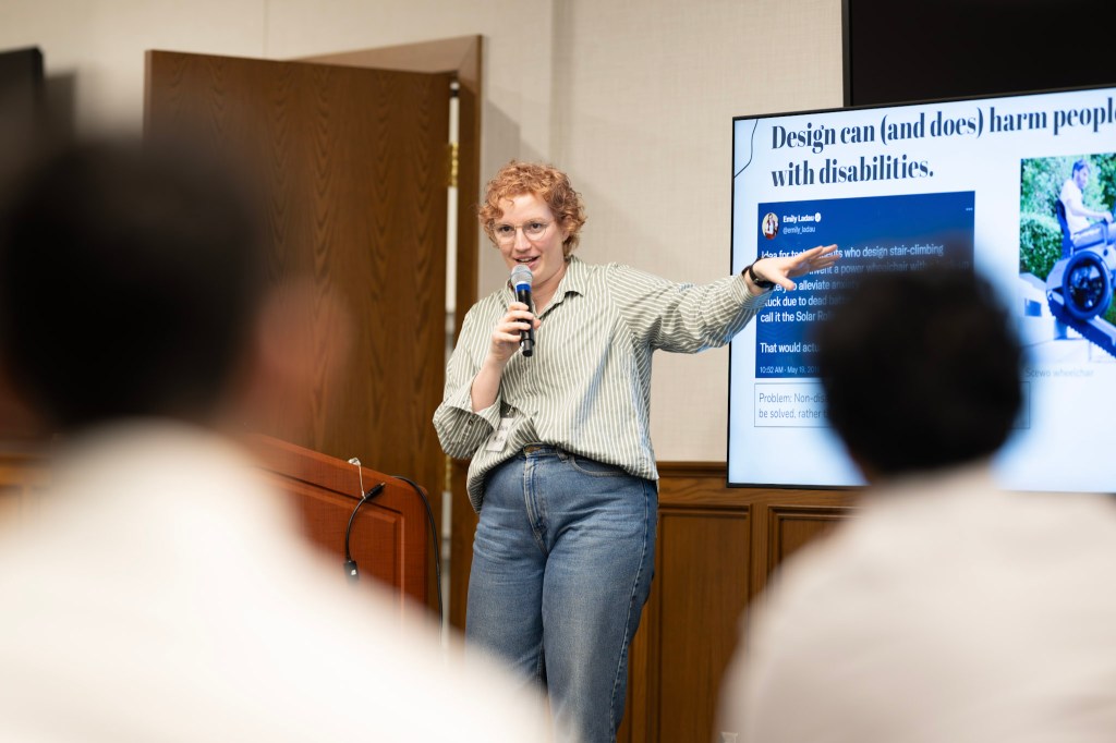 Laura is standing in front of a room full of professionals enthusiastically gesturing at her slides which say "Design can and does harm disabled people."